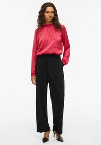 Blusa rossa in satin con collo e maniche arricciati, abbinata a pantaloni neri a vita alta. Il modello indossa tacchi a spillo neri. Sfondo semplice.