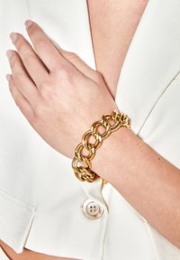 Bracciale in metallo dorato con maglie intrecciate, caratterizzato da una finitura liscia e lucida. Indossato su un polso su uno sfondo di indumento bianco.