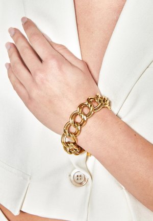 Gold metal chain bracelet with interlocking links, featuring a smooth, polished finish. Worn on a wrist against a white garment background.