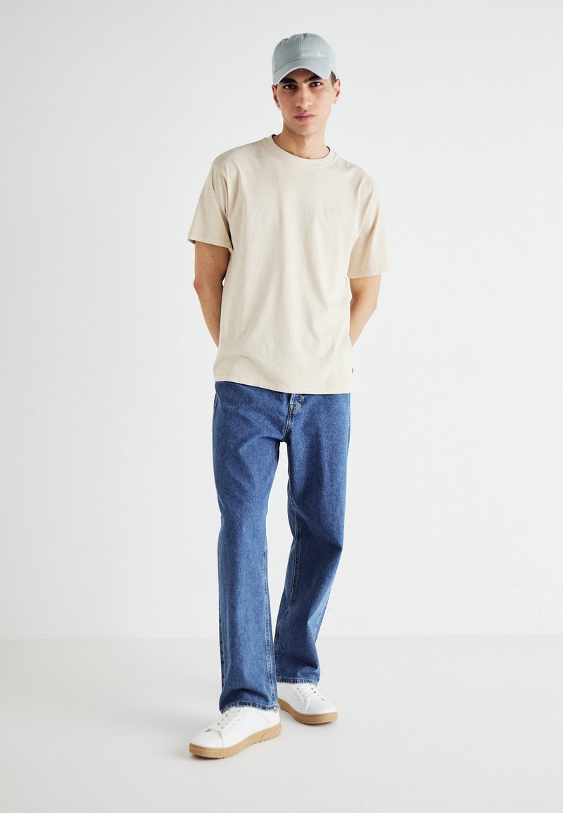 T-shirt in cotone beige, vestibilità comoda, abbinata a jeans in denim blu chiaro. Sneakers bianche con suole beige. Un cappellino grigio completa il look casual.