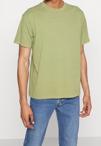 T-shirt en coton vert olive avec un col rond et des manches courtes. Présente un petit logo sur la poitrine. Design à coupe classique.