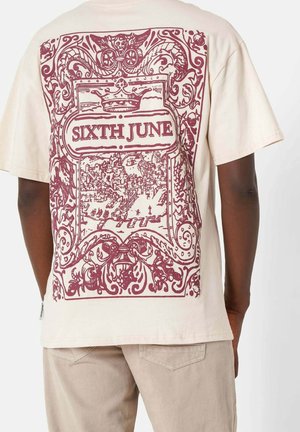 Beiges T-Shirt mit burgunderfarbenem, filigranem verziertem Rahmen, einer Krone, einer Stadtsilhouette und dem Text „SIXTH JUNE“ auf dem Rücken, getragen von einer stehenden Person.