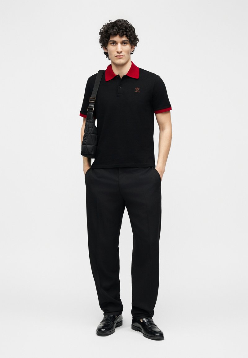 Young man standing with hands in pockets, wearing black pants, black polo shirt with red collar and cuffs, and black shoulder bag.