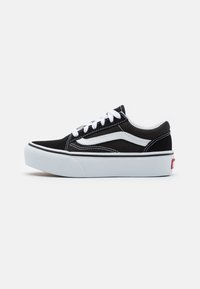 Zapatillas de lona negras con cordones blancos y una franja lateral blanca. Presentan una suela de goma blanca gruesa y una etiqueta de logo roja en el talón.