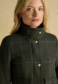 Giacca verde in misto lana con colletto alto, bottoni dorati e un motivo a quadri con linee di verde scuro, verde chiaro e viola.