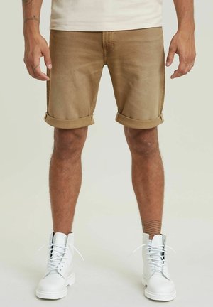 Homme portant un short marron retroussé, des bottes blanches à lacets et une chemise blanche, se tenant devant un fond clair uni.