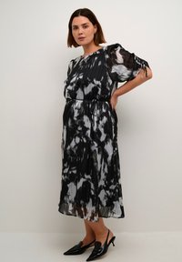 Zwart-witte plissé midi-jurk met abstract patroon, korte pofmouwen en een trekkoord in de taille. Gemaakt met puntige zwarte hakken.