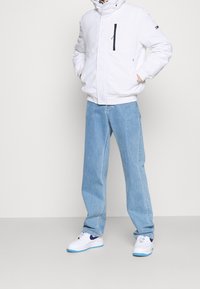 Hvid fleecejakke med en frontlynlås lomme, parret med lyseblå denim bukser og hvide sneakers med blå detaljer.