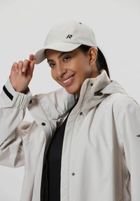 Vit keps med svart logotyp, ljusfärgad vattentät jacka, svart strukturerad topp under, mjukt tyg och kontrasterande ränder på ärmarna.