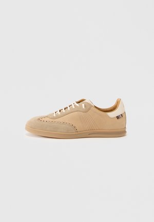 Beige suede sneaker with perforated detailing, cream laces, and a textured tan sole, featuring a color-contrasting accent on the heel.