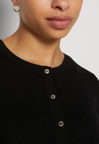 Zwarte cardigan met een ronde halslijn, voorzien van drie gouden drukknoopjes en een geribbelde textuur. De stof lijkt zacht en aansluitend.