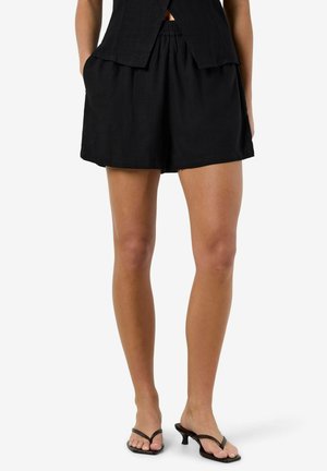 Nő laza fekete rövidnadrágban, oldalán zsebekkel, fekete magassarkú papucsokkal, fehér háttér előtt áll.