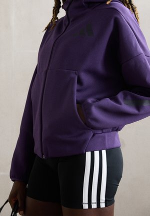 Osoba w fioletowej bluzie na suwak z ręką w kieszeni oraz czarnych szortach z trzema białymi paskami po bokach.