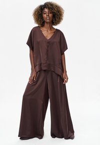 Bruine satijnen top met V-hals en korte mouwen, gecombineerd met wijde broekspijpen in dezelfde bruine satijnstof. Gladde textuur en ontspannen silhouet overal.