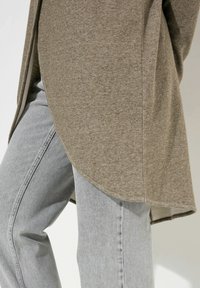 Lungo maglione beige a lavorazione a maglia con orlo arrotondato, abbinato a jeans grigi chiari. Il maglione presenta un tessuto testurizzato e una vestibilità rilassata.