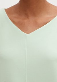 Miętowa bluzka z dekoltem w kształcie V, wykonana z miękkiego, gładkiego materiału. Posiada centralny szew biegnący pionowo wzdłuż przodu, co nadaje jej elegancki wygląd.