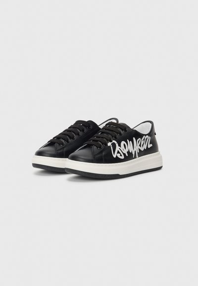 Baskets en cuir noir avec semelle en caoutchouc blanc, arborant un texte graphique blanc sur le côté et des lacets noirs. Texture lisse, design décontracté.