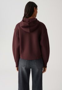 Bordeaux gekorte hoodie met een capuchon, brede mouwen en een gladde textuur. Gecombineerd met hoog-waist donkergrijze jeans met achterzakken.