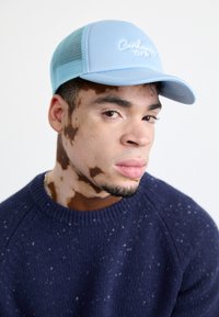 Gorra de malla azul claro con logo bordado, llevada con un suéter oscuro de color marino con textura moteada. Primer plano del rostro de un joven.