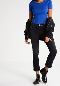T-shirt blu aderente, jeans neri cropped e stivaletti neri lucidi. Giacca nera appoggiata su un braccio. Design semplice, senza motivi visibili.