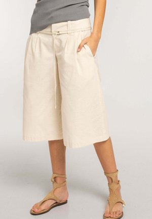 Persona que lleva pantalones cortos beige de pierna ancha hasta la rodilla con bolsillos, una camiseta sin mangas gris y sandalias planas marrones con tiras.