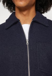Navy wool blend jacket features a zip closure, pointed collar, and a patch pocket on the chest with a soft, textured finish.