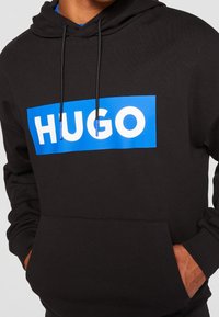 Sweat à capuche noir avec un logo rectangulaire bleu affichant le texte blanc "HUGO," porté par une personne les mains dans la poche avant.