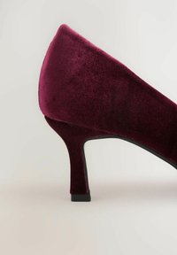 Primer plano de un zapato de salón de terciopelo burdeos con tacón medio, parte trasera redondeada y suela negra sobre un fondo claro.