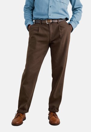 Mand iført brune plisserede bukser, brun læderbælte, brune lædersko og en lyseblå skjorte med knapper med hænderne i lommerne.