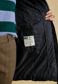 Wnętrze czarnej puchowej kurtki z etykietą marki "JOULES". Widoczne spodnie w kratę z brązowymi, beżowymi i czerwonymi wzorami.