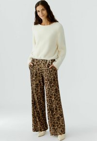 Vrouw staat met haar handen in haar zakken, draagt een witte fluffy trui, brede broek met luipaardprint en crèmekleurige puntige schoenen, tegen een effen achtergrond.