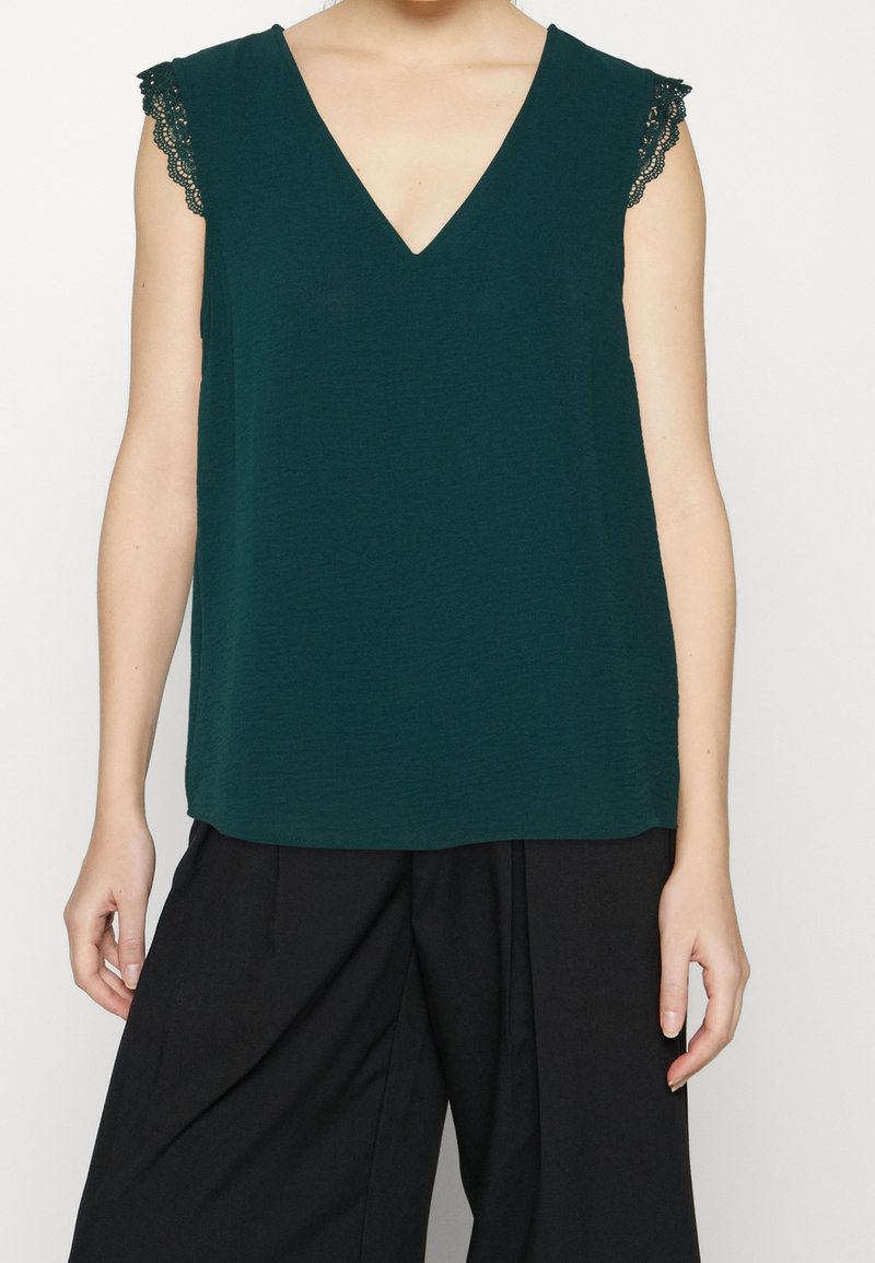 Dark green sleeveless top with a v-neck and lace trim on shoulders, featuring a textured fabric and a loose fit. Paired with black pants.