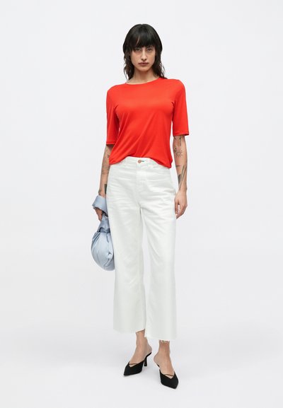 Piros rövid ujjú póló, fehér bő szárú nadrág, világoskék felsőt tartva. Fekete magas sarkú cipő. Minimalista és modern design.