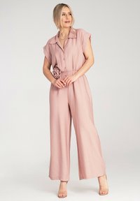 Różowy kombinezon z krótkimi rękawami, zasuwaną z przodu na guziki, szerokimi nogawkami oraz materiałowym kwiatem na talii. Gładka tekstura, lekki materiał.