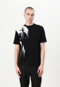 Črna bombažna majica s kratkimi rokavi, ki na levi strani prikazuje grafični potisk v beli barvi, ki prikazuje abstraktne oblike in linije. Standarden kroj s trupom in okroglim izrezom.