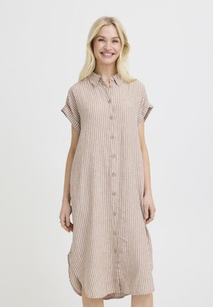 Naeratav naine, kes kannab põlveni ulatuvat beežikat kleiti valgete vertikaalsete triipude ja nuppudega, seistes ühevärvilise valge tausta ees.