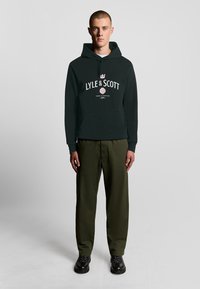 Felpa verde scuro con tasca frontale e testo del logo bianco. Abbinata a pantaloni verde oliva e stivali neri con lacci. Design semplice e casual.