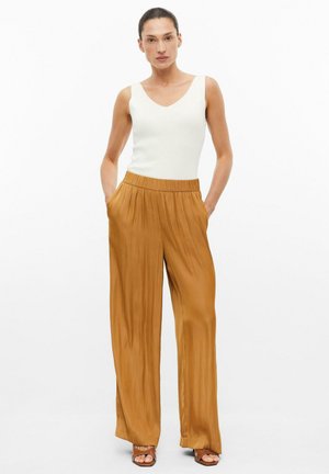 Femme debout face à l'avant, portant un haut blanc sans manches, un pantalon large en satin bronze et des sandales à talons, les mains dans les poches.