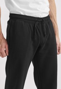 Svarta bomulls-joggers med resår i midjan och dragsko, med sidofickor och avslappnad passform. Mjuk textur med minimal detaljering.