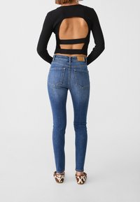 Top negro de manga larga acanalado con espalda descubierta y detalles de tiras, combinado con jeans ajustados azules que cuentan con dos bolsillos traseros y una etiqueta marrón.