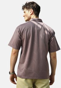 Koszulka polo z krótkim rękawem w stonowanym mauve, z teksturowanym wzorem, klasycznym kołnierzykiem i gładkim wykończeniem materiału, noszona z jasnymi spodniami.