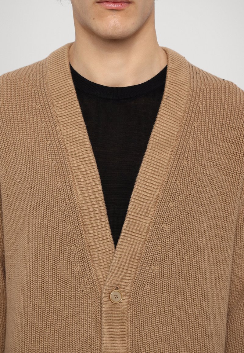 Beige gestrickte Strickjacke mit tiefem V-Ausschnitt, geripptem Kragen und einer Knopfverschluss. Sie verfügt über einen strukturierten Stoff und ein kontrastierendes schwarzes Innenhemd, das sichtbar ist.