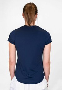 Donna con coda di cavallo castana che indossa una maglietta blu navy a maniche corte e una gonna bianca, in piedi con le spalle alla macchina fotografica.