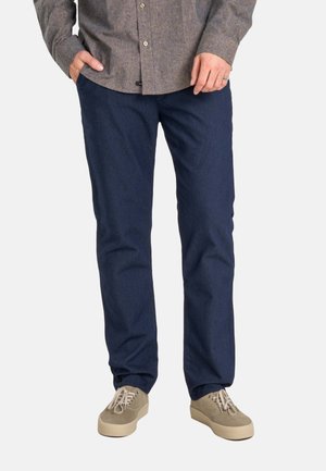 Man wearing dark blue pants, beige sneakers, and a brown button-up shirt with hands in pockets against a white background.