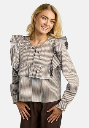 Lächelnde Frau mit blondem Haar trägt eine beige-weiß gestreifte Bluse mit Rüschen an den Schultern und einer Krawattenschleife am Ausschnitt, posiert mit den Händen in die Hüften gestemmt.