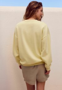 Helles gelbes Sweatshirt mit überschnittenen Schultern und gerippten Bündchen, kombiniert mit strukturierten beigen Shorts, vor einem neutralen Hintergrund.