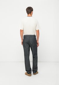 Hombre de espaldas luciendo una camisa blanca de manga corta, pantalones de rayas gris oscuro y zapatos casuales color beige, contra un fondo blanco.