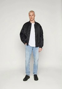 Chaqueta bomber negra con una textura suave, combinada con una camiseta blanca, jeans de color azul claro y zapatillas negras. Diseño y ajuste sencillos.