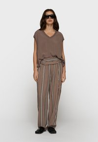 Mujer con top marrón de cuello en V oversize, pantalones de rayas anchas de cintura alta con cinturón, zapatos negros y grandes gafas de sol negras, de pie frente a un fondo blanco.