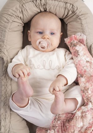 Baby liegt in einem beigen gepolsterten Nest, trägt ein cremefarbenes Outfit mit dem Schriftzug "hello", hält die Füße, Schnuller im Mund, mit einer rosa Blumen-Decke in der Nähe.